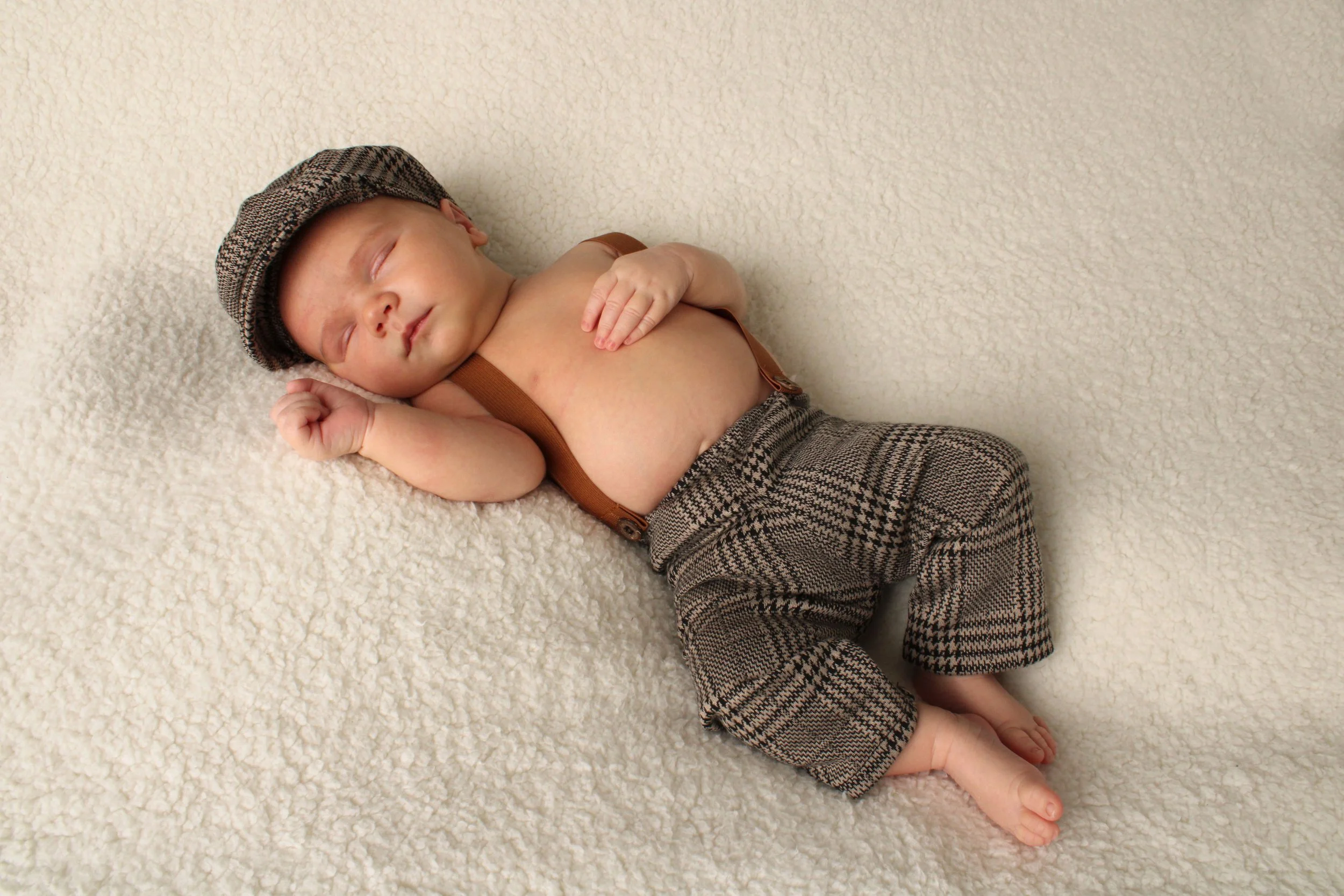 Sleeping newborn baby boy wearing a houndstooth cap and suspenders during a Nassau County newborn photography session